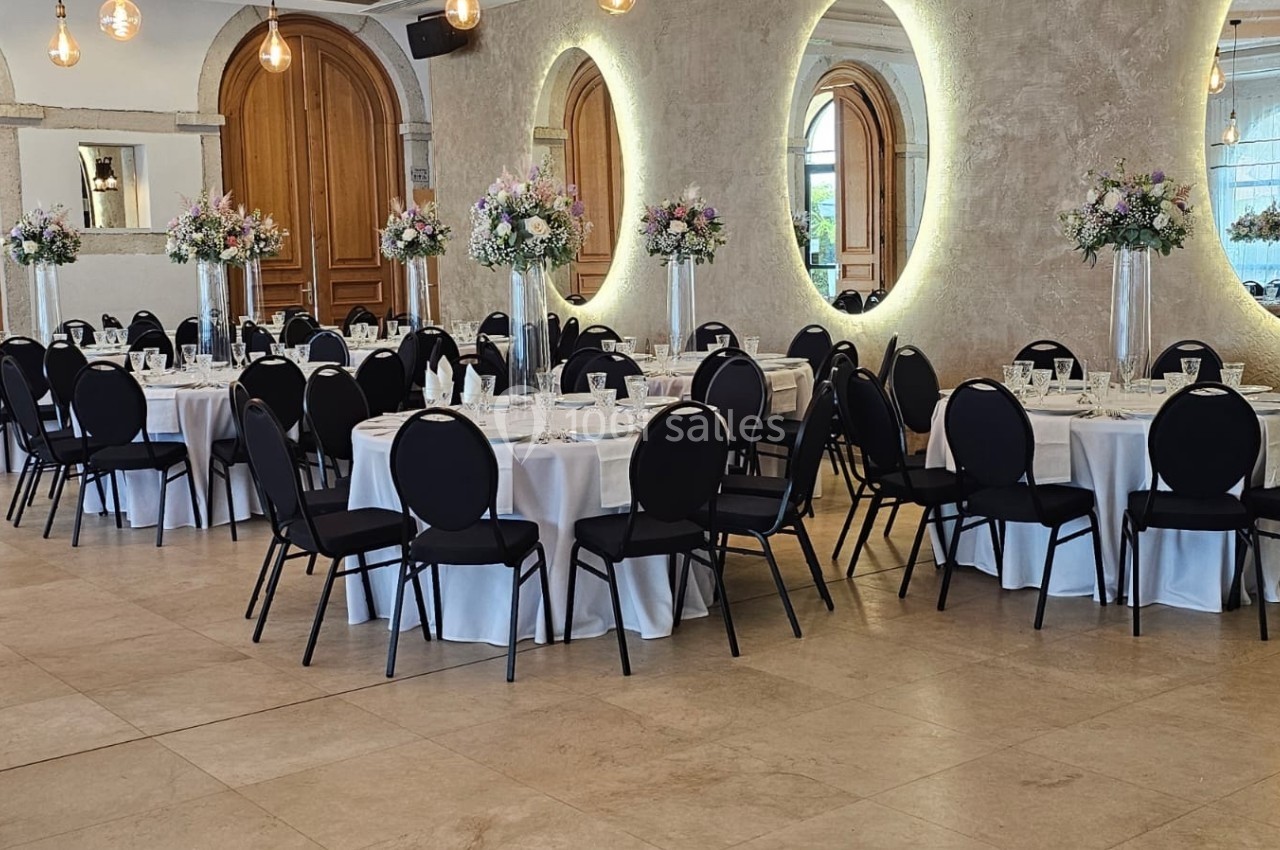 Salle de réception élégante avec tables rondes dressées, chaises noires et décorations florales sur de hauts vases en verre.