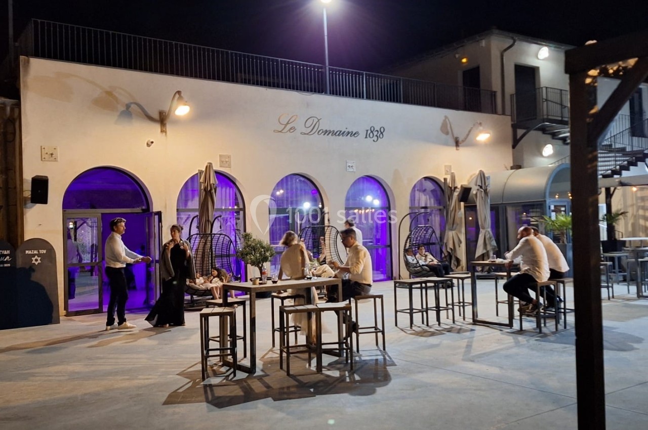 Terrasse éclairée en soirée avec des tables hautes, des invités debout et assis, devant un bâtiment moderne.