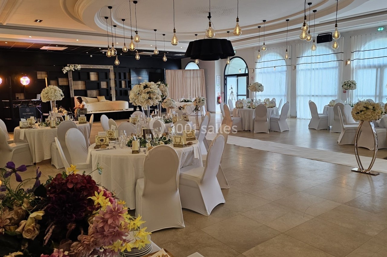Salle de réception élégante avec tables rondes décorées de fleurs blanches, chaises blanches et éclairage suspendu.