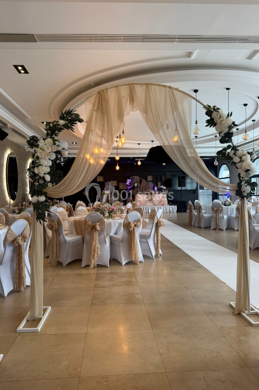 Salle de réception décorée avec des tables dressées, des chaises ornées de nœuds beige et une arche florale en premier plan.