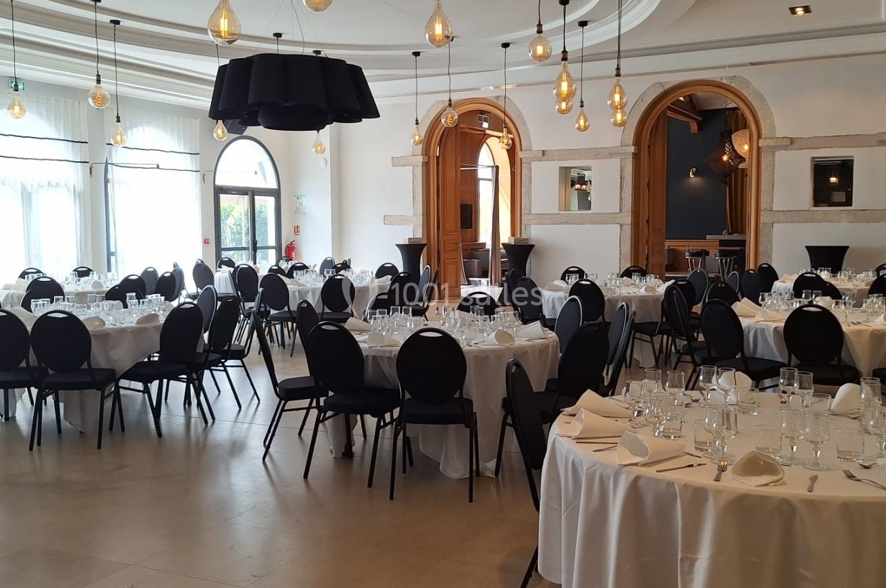 Salle de réception lumineuse avec tables rondes dressées, chaises noires et décorations suspendues modernes.