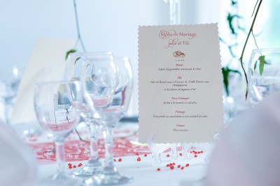 Carte de menu de mariage posée sur une table décorée avec des verres, pétales rouges et nappes blanches.