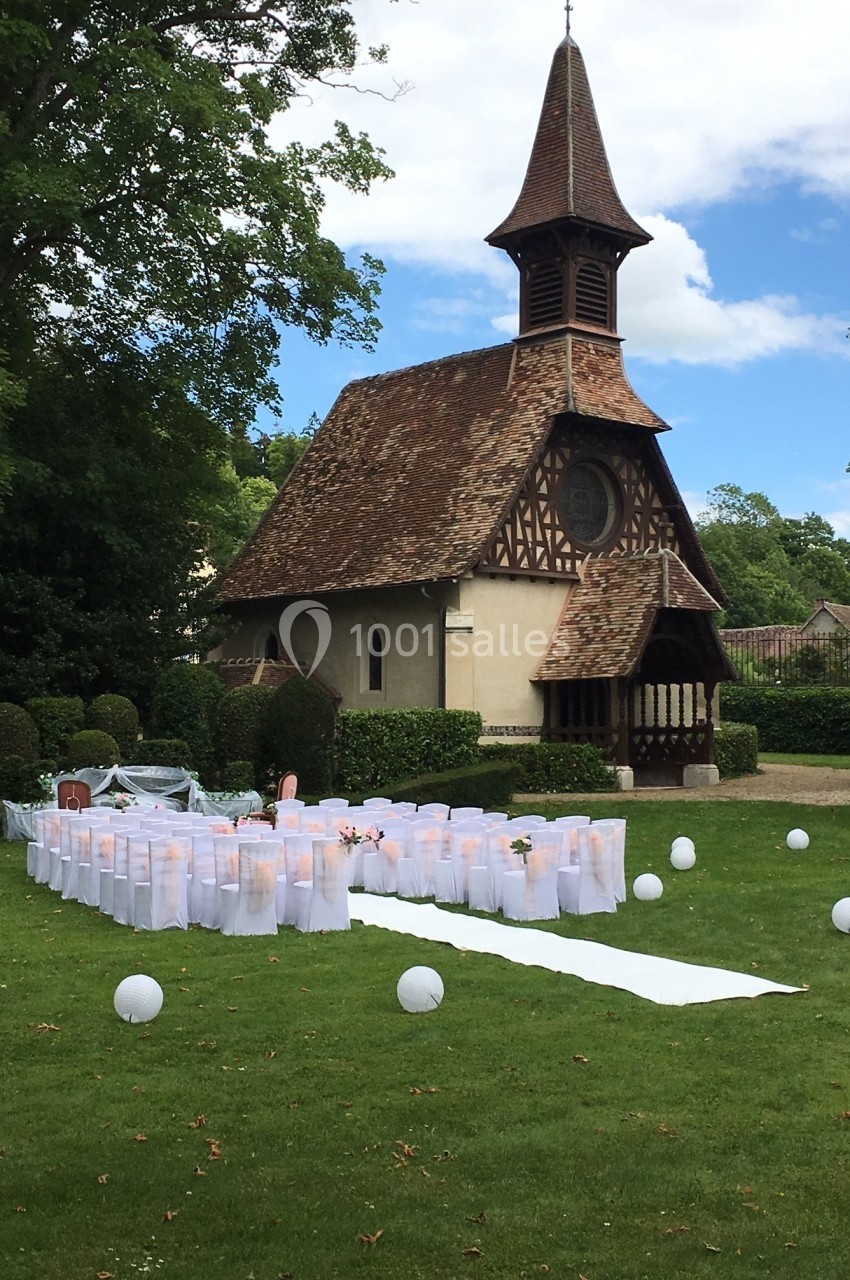 Allée blanche et chaises décorées pour une cérémonie en plein air devant une petite chapelle entourée de verdure.