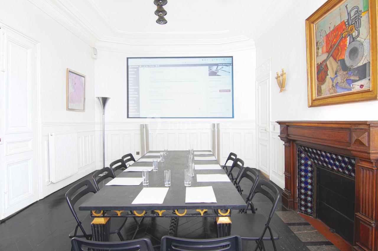 Salle de réunion lumineuse avec une grande table, des chaises noires, un écran mural et une cheminée décorative.