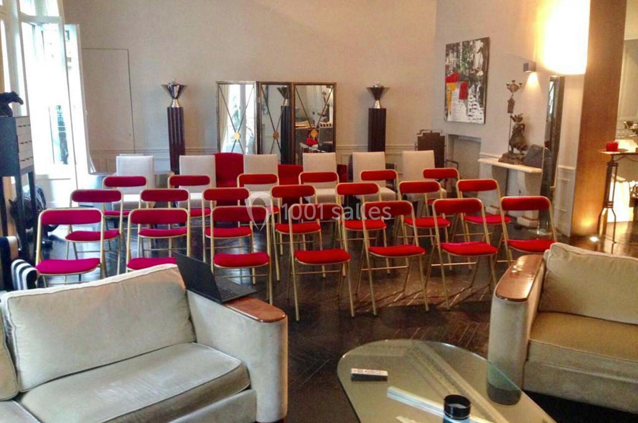 Salle aménagée avec des rangées de chaises rouges face à un espace de présentation, entourée de meubles et de décorations.