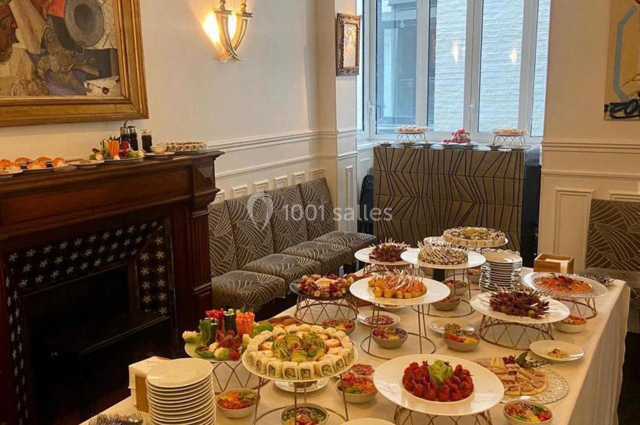 Buffet varié avec plats et desserts disposés sur des présentoirs dans une salle lumineuse avec cheminée et tableaux.