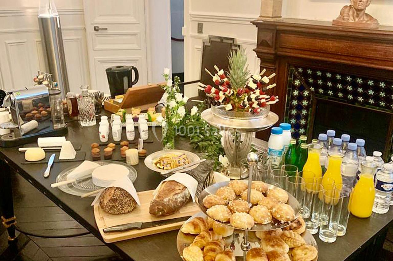 Table garnie de viennoiseries, pains, jus, boissons et fruits, disposée dans une salle élégante avec cheminée en arrière…