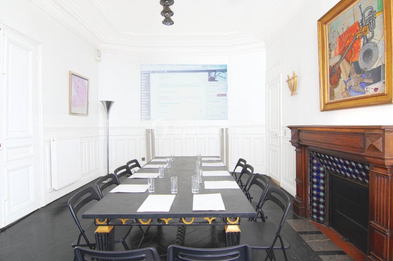 Salle de réunion lumineuse avec une grande table, des chaises noires, un écran projeté et une cheminée décorative.