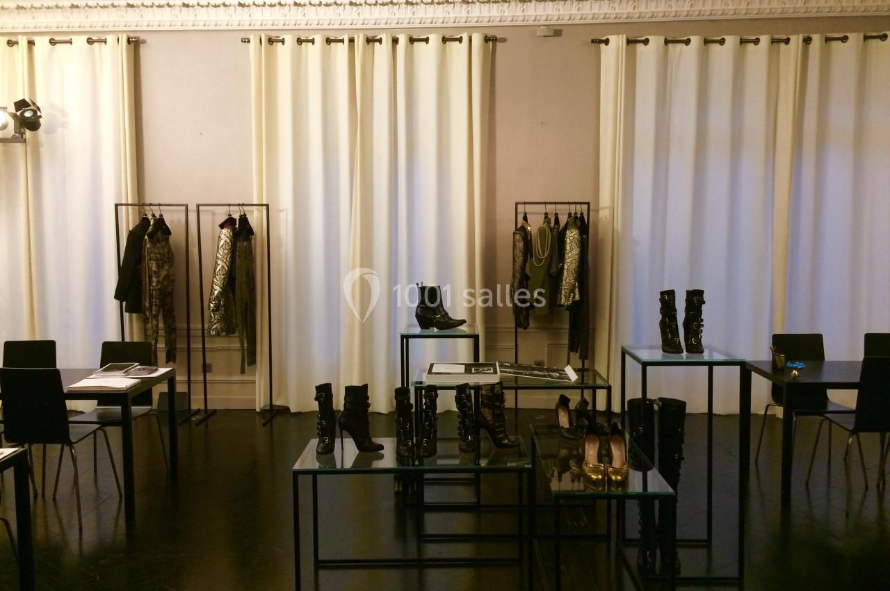 Salle d'exposition avec vêtements suspendus, bottes et chaussures exposées sur des tables en verre noir.