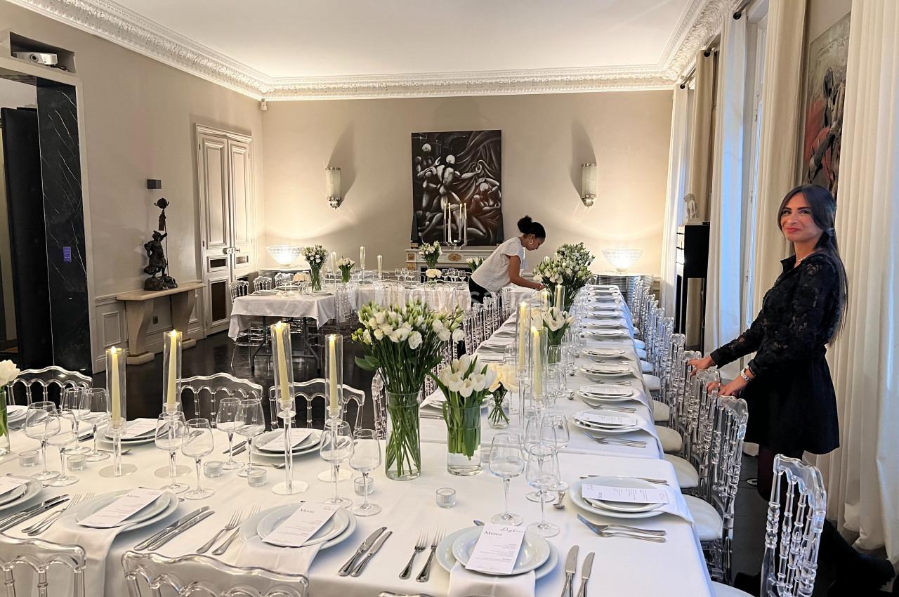 Salle élégante avec une longue table dressée pour un dîner, décorée de fleurs blanches et bougies, avec deux personnes…