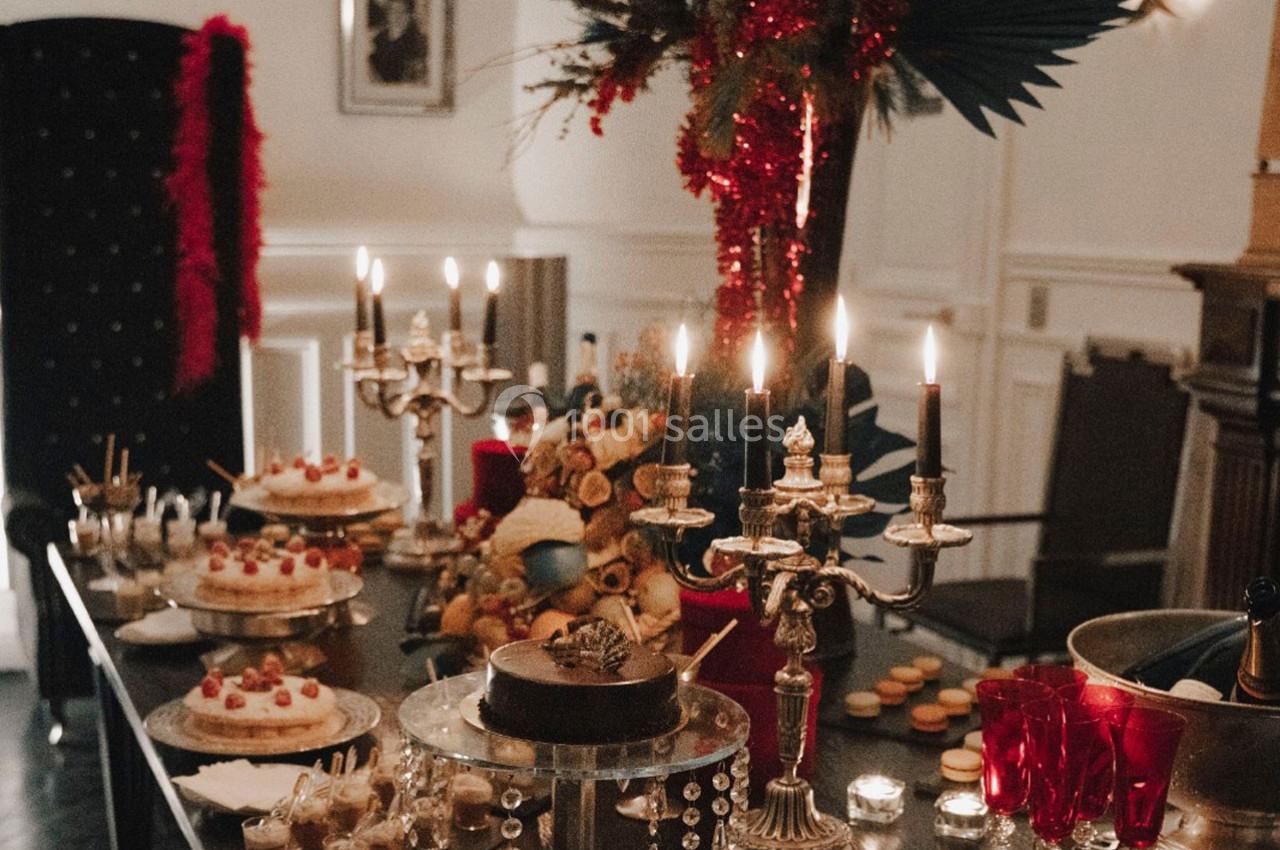 Table festive décorée avec chandeliers, desserts variés, verres rouges et ornements rouges dans un intérieur élégant.
