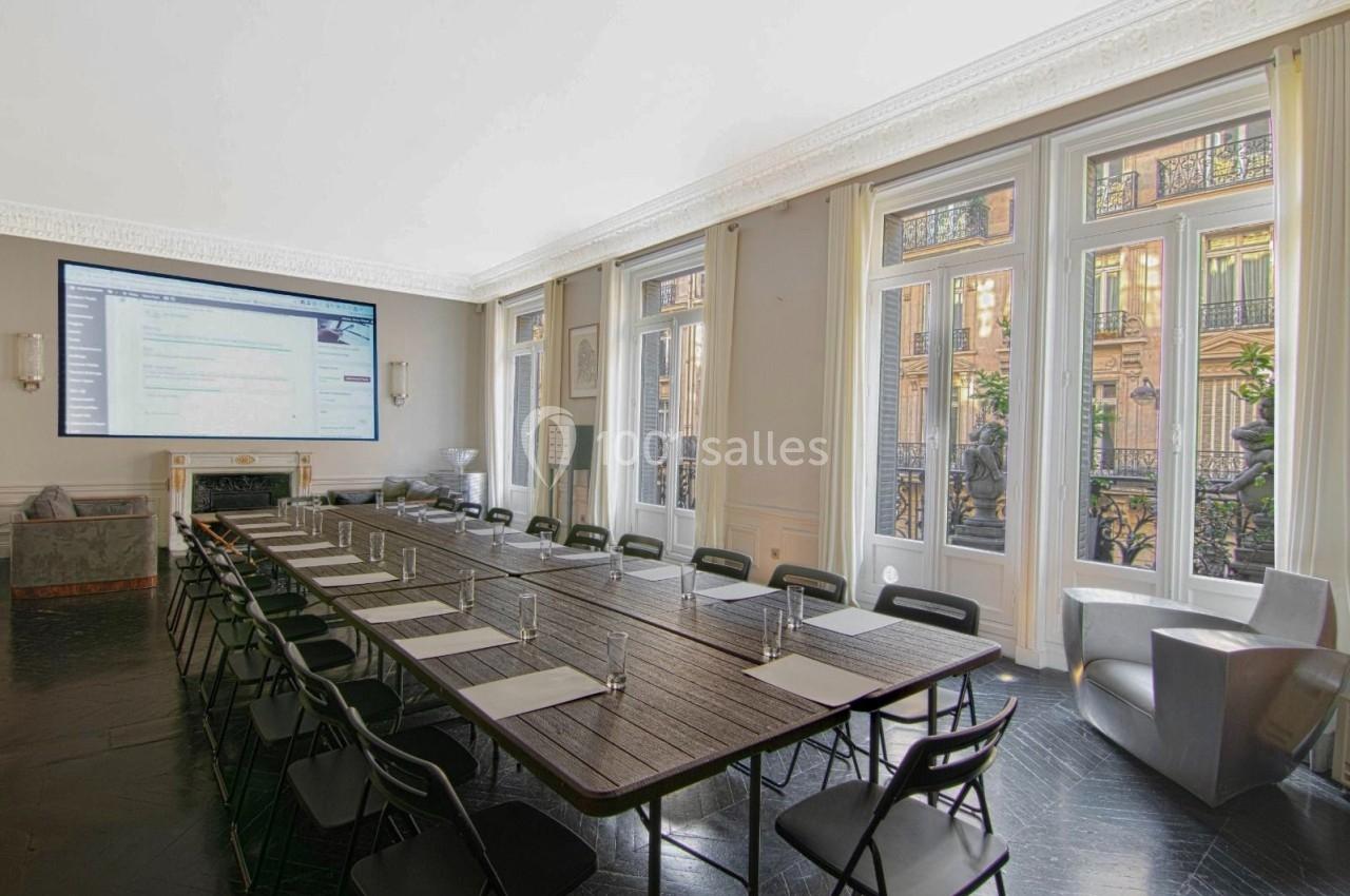 Salle de réunion lumineuse avec une grande table en bois, chaises pliantes, écran mural et fenêtres donnant sur un balcon.