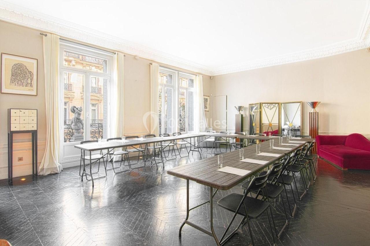 Salle lumineuse avec de grandes fenêtres, tables alignées en U, chaises pliantes et décorations élégantes.