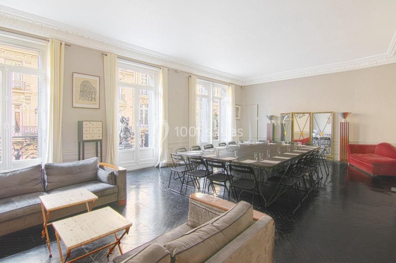 Salle lumineuse avec grandes fenêtres, parquet sombre, table de réunion, chaises pliantes et mobilier de salon.