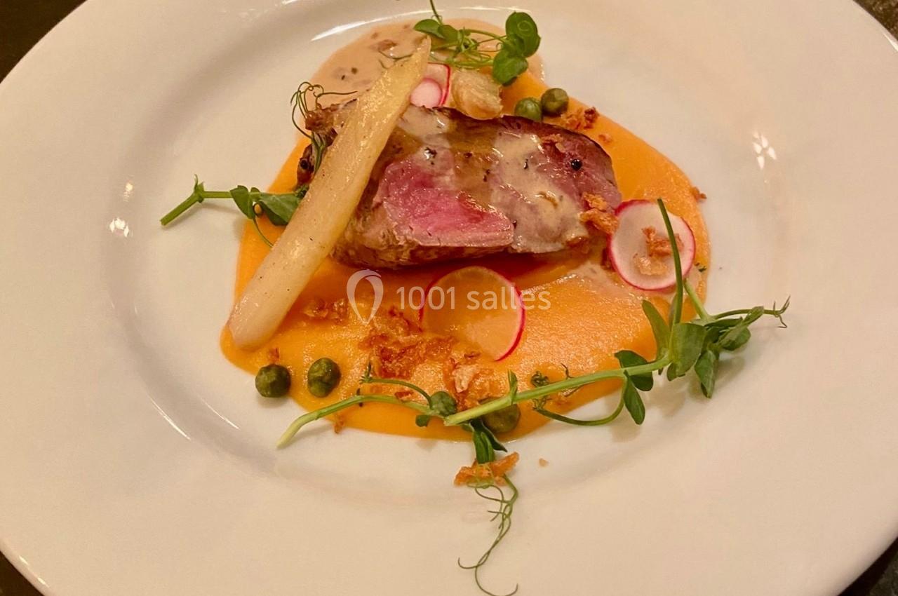 Assiette gastronomique avec viande, purée orange, asperge blanche, radis, câpres et herbes fraîches sur une table sombre.