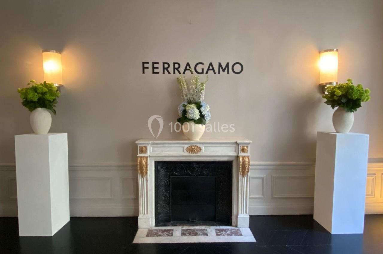 Cheminée blanche décorative encadrée de deux colonnes avec vases fleuris, sous le logo ’Ferragamo’ sur un mur beige.