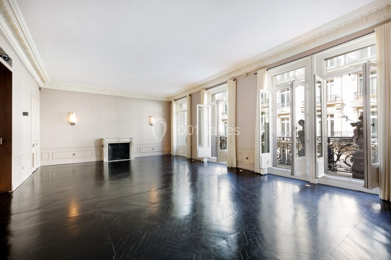 Salon vide avec parquet sombre, cheminée, moulures au plafond et grandes fenêtres donnant sur un balcon en fer forgé.