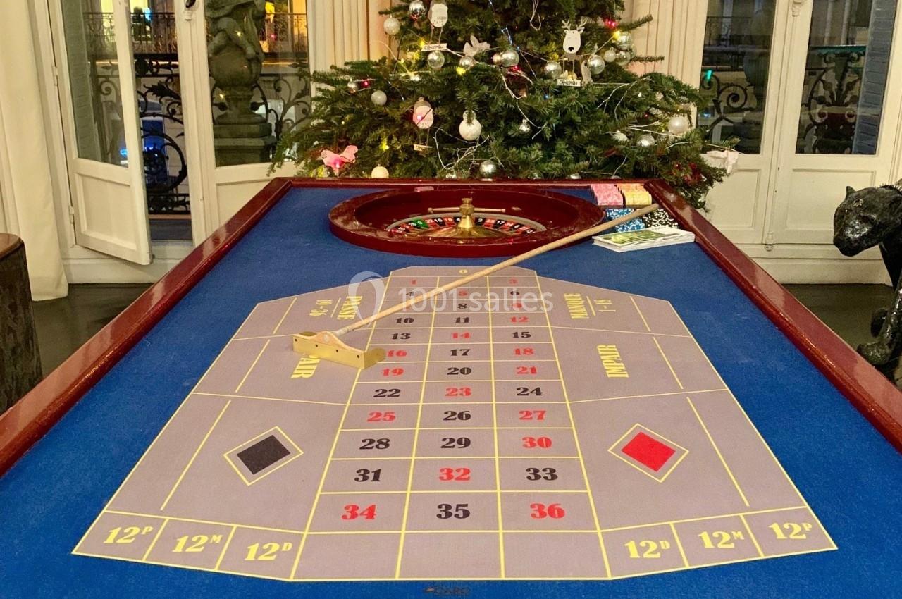 Table de roulette avec tapis de jeu bleu, devant un sapin de Noël décoré, dans une pièce avec fenêtres vitrées.