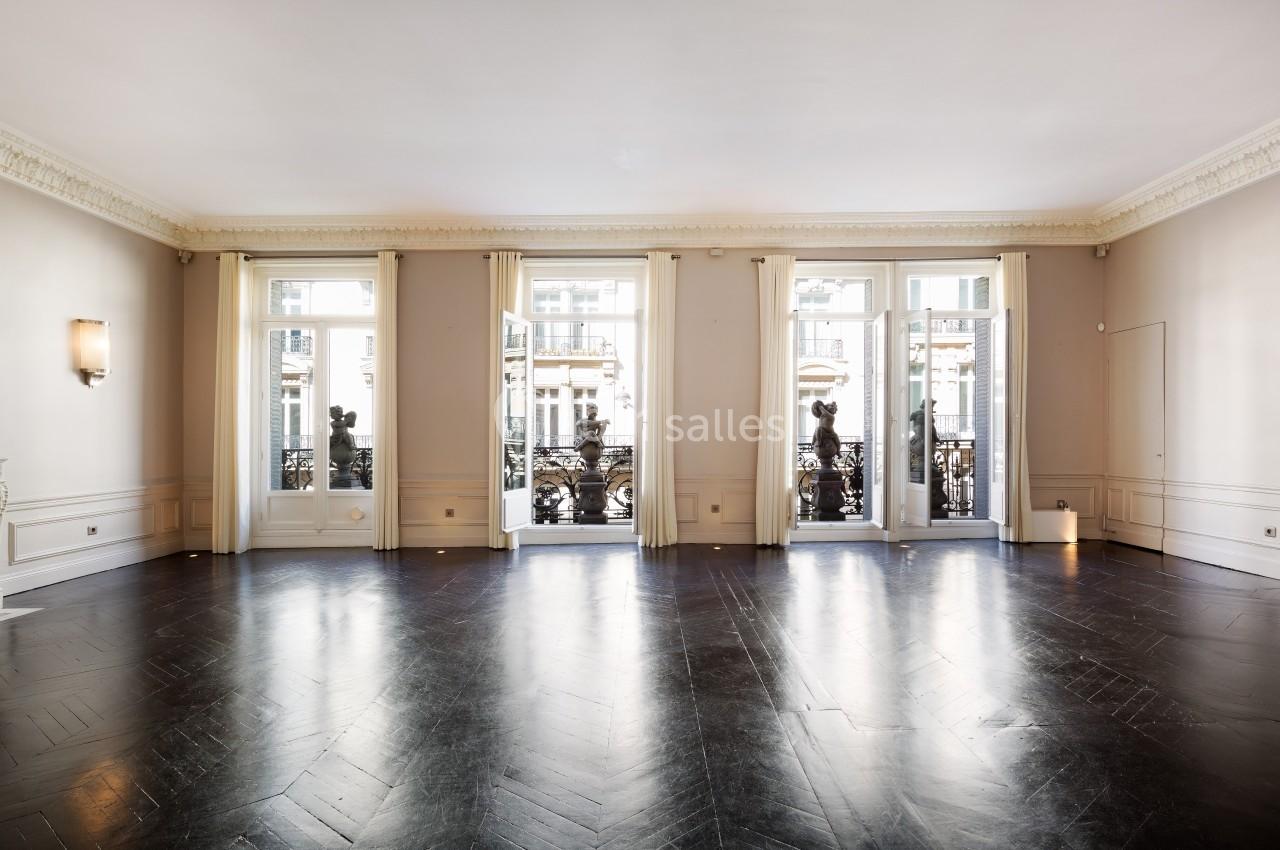 Salle lumineuse avec parquet sombre, moulures au plafond et grandes fenêtres donnant sur un balcon en fer forgé.