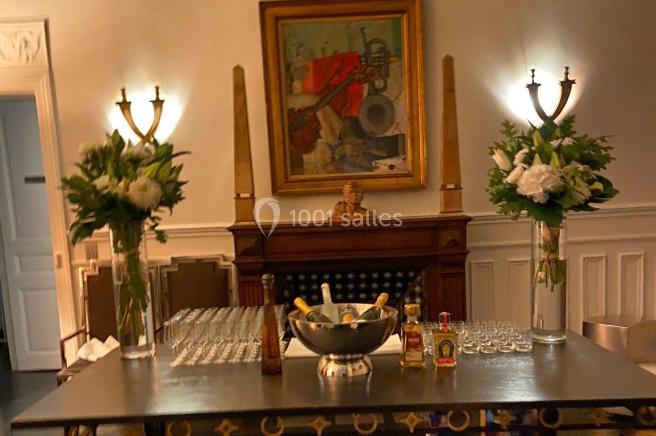 Table avec seau à champagne, verres alignés, fleurs blanches en vase, devant une cheminée ornée d'un tableau coloré.