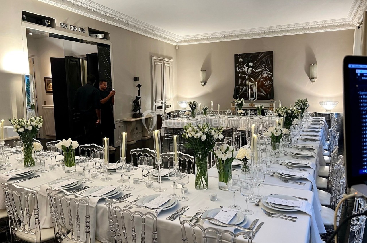 Salle élégante avec tables longues dressées, décorées de bouquets de fleurs blanches et de bougies, prête pour un événement.