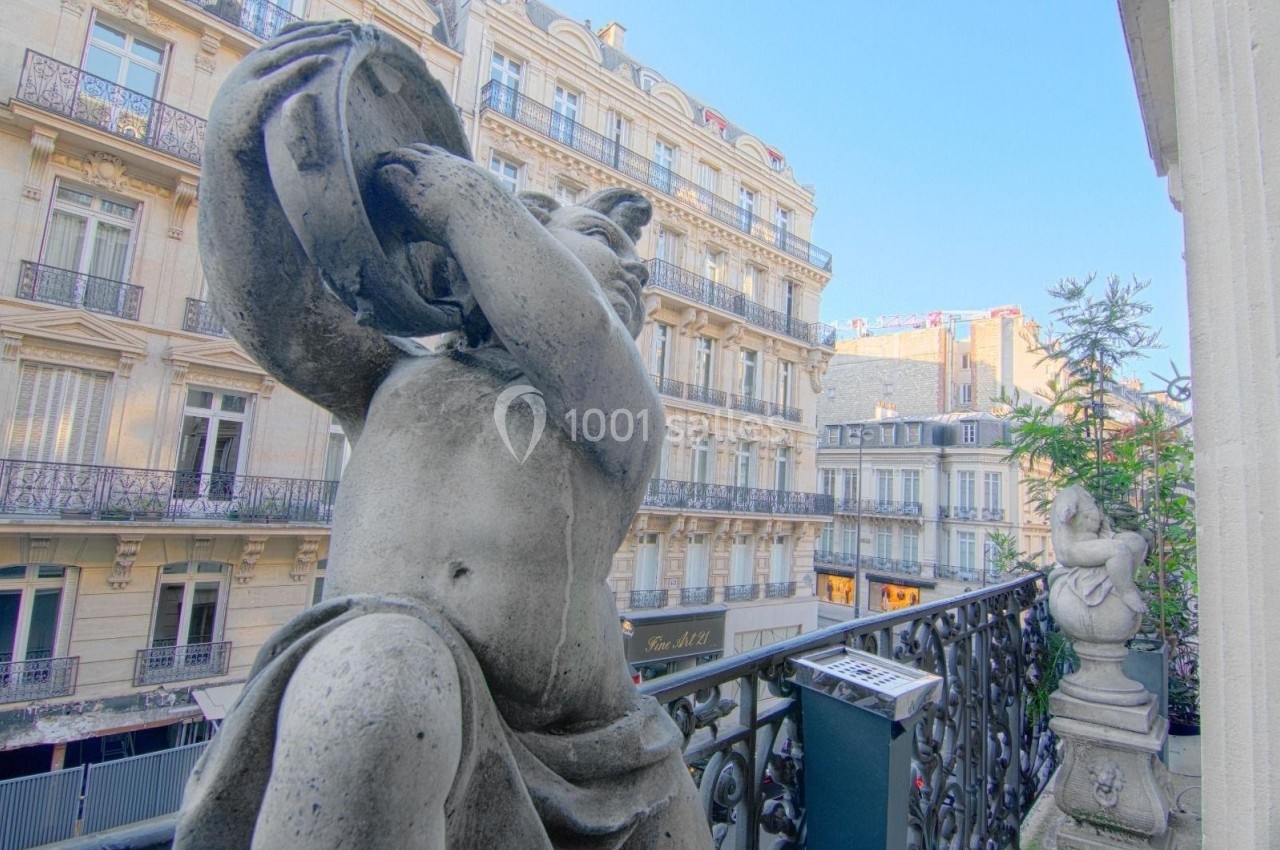Statue en pierre sur un balcon décoré, avec des immeubles haussmanniens en arrière-plan sous un ciel dégagé.