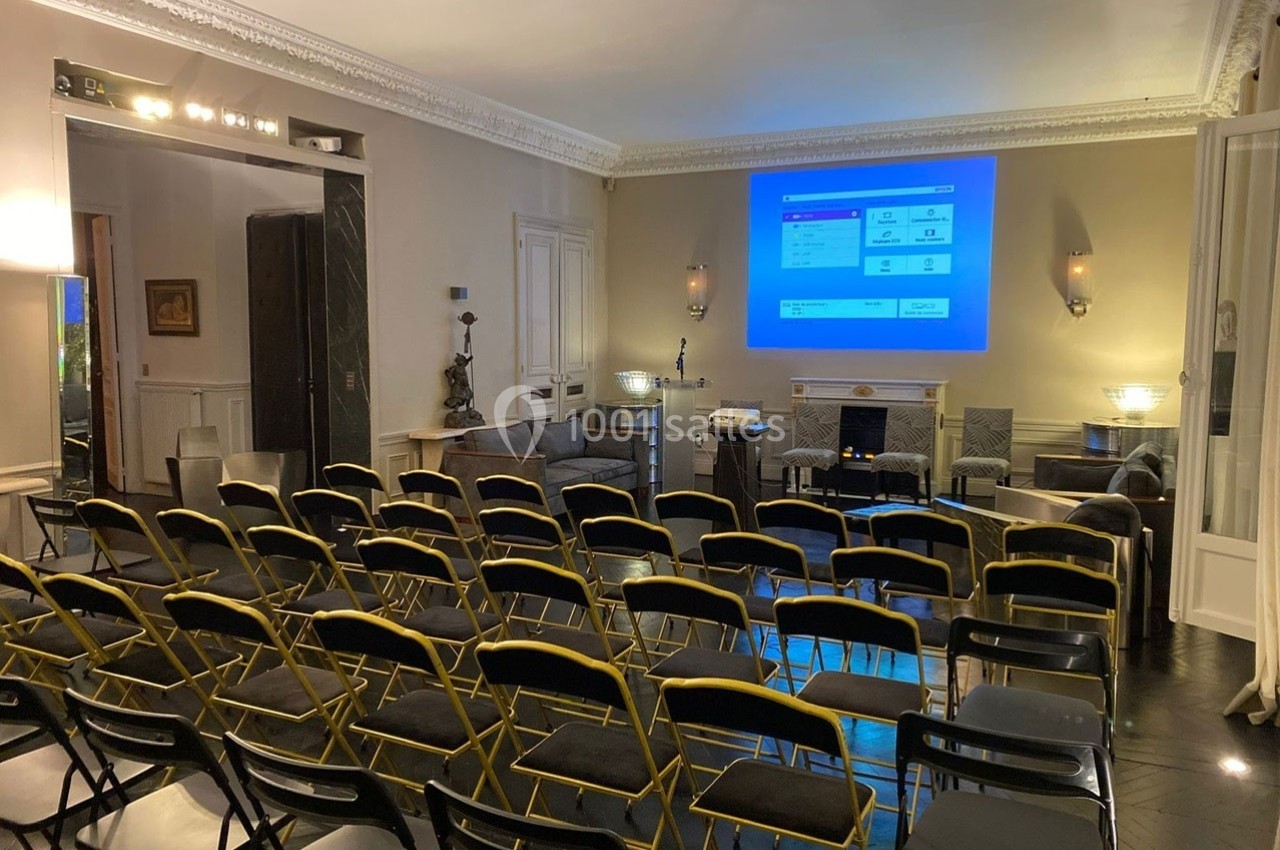 Salle de conférence avec des rangées de chaises noires face à un écran de projection allumé sur un mur beige.