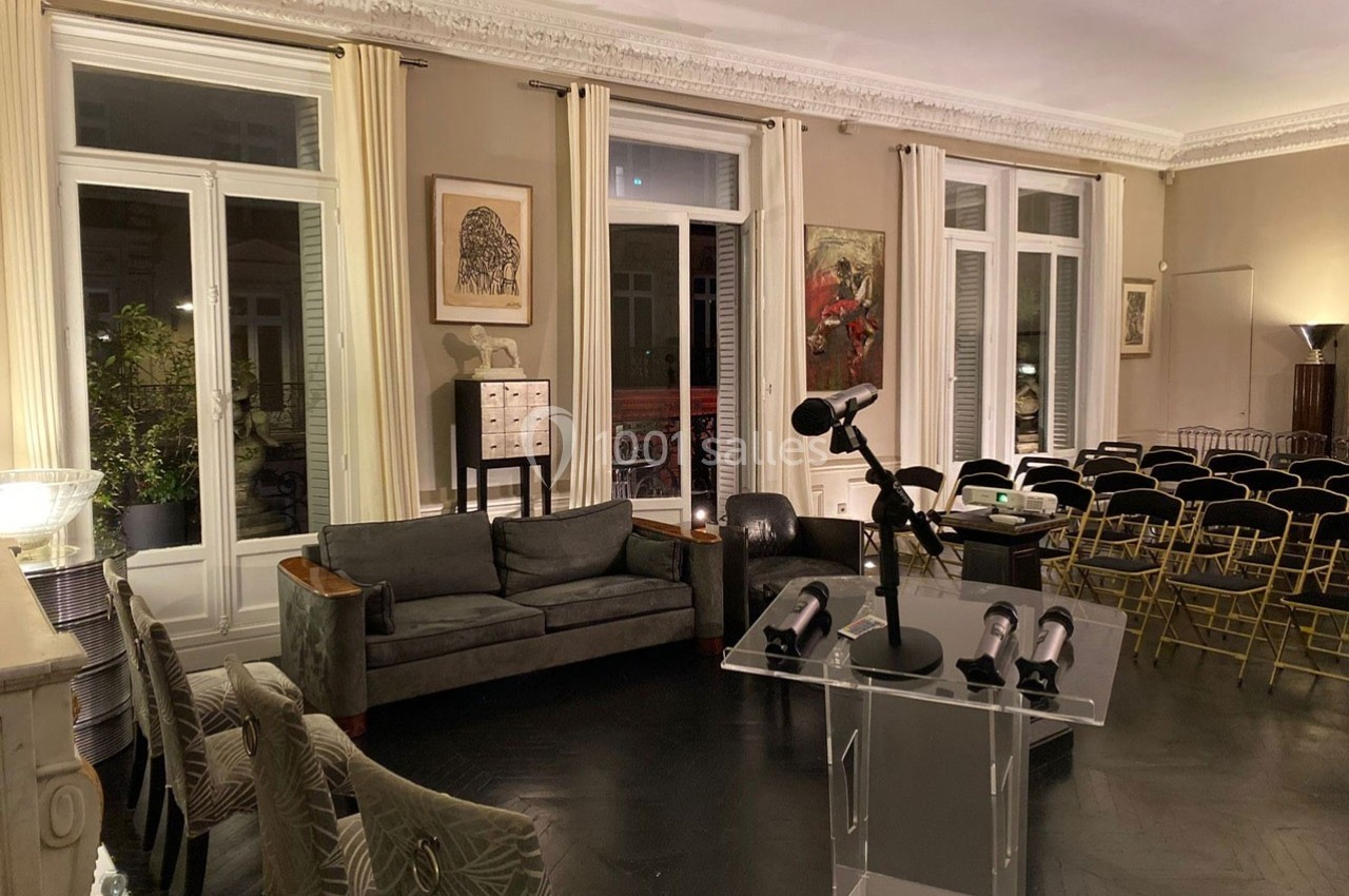 Salle élégante avec canapé, chaises alignées, pupitre transparent et grandes fenêtres avec rideaux clairs.