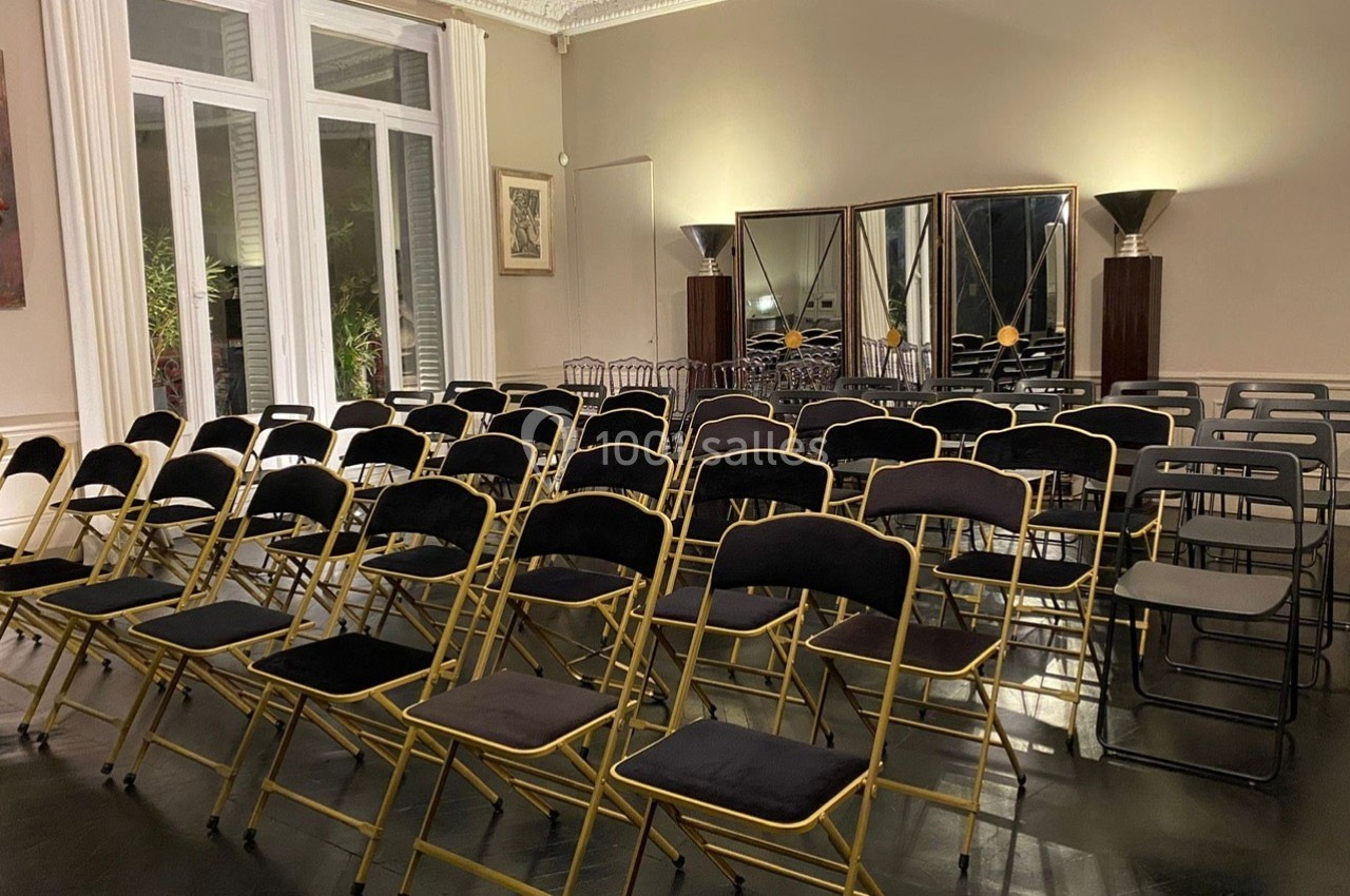 Chaises pliantes noires et dorées alignées dans une salle lumineuse avec miroirs et grandes fenêtres.
