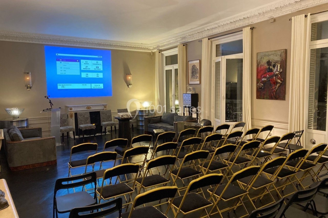 Salle de conférence avec des chaises alignées face à un écran de projection allumé, dans un cadre intérieur élégant.