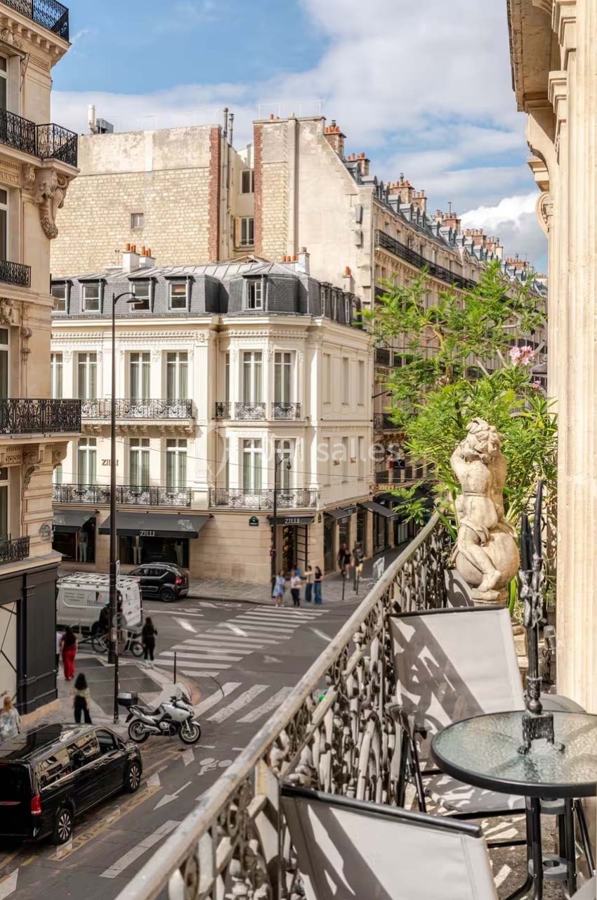 Vue d'une rue parisienne depuis un balcon, avec des immeubles haussmanniens, des passants et des véhicules.