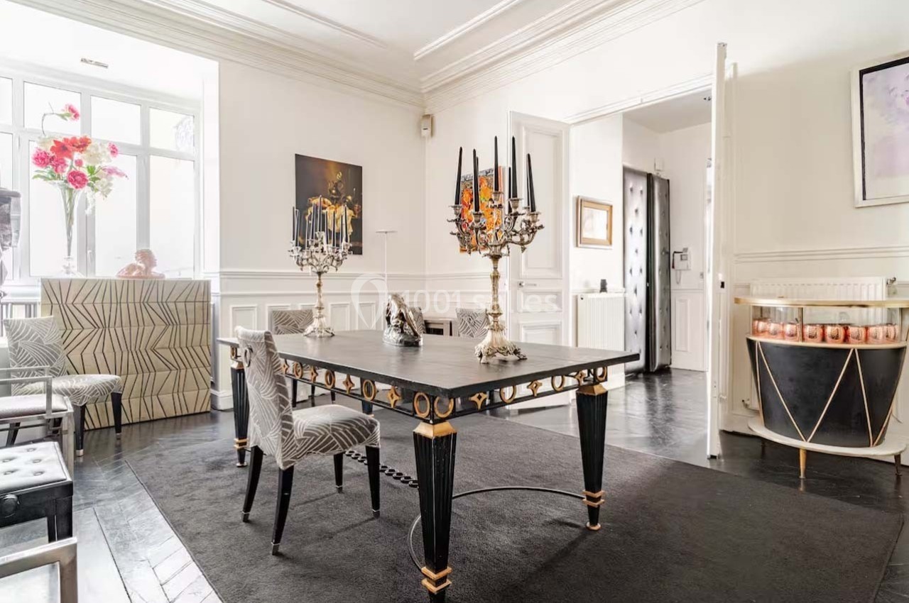 Salle à manger élégante avec table noire ornée, chandeliers, chaises à motifs et lumière naturelle provenant d'une fenêtre.