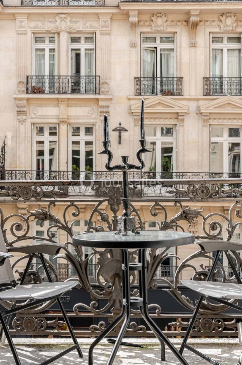 Table ronde en métal noir avec chandelier et chaises, placée sur un balcon avec vue sur une façade d'immeuble haussmannien.