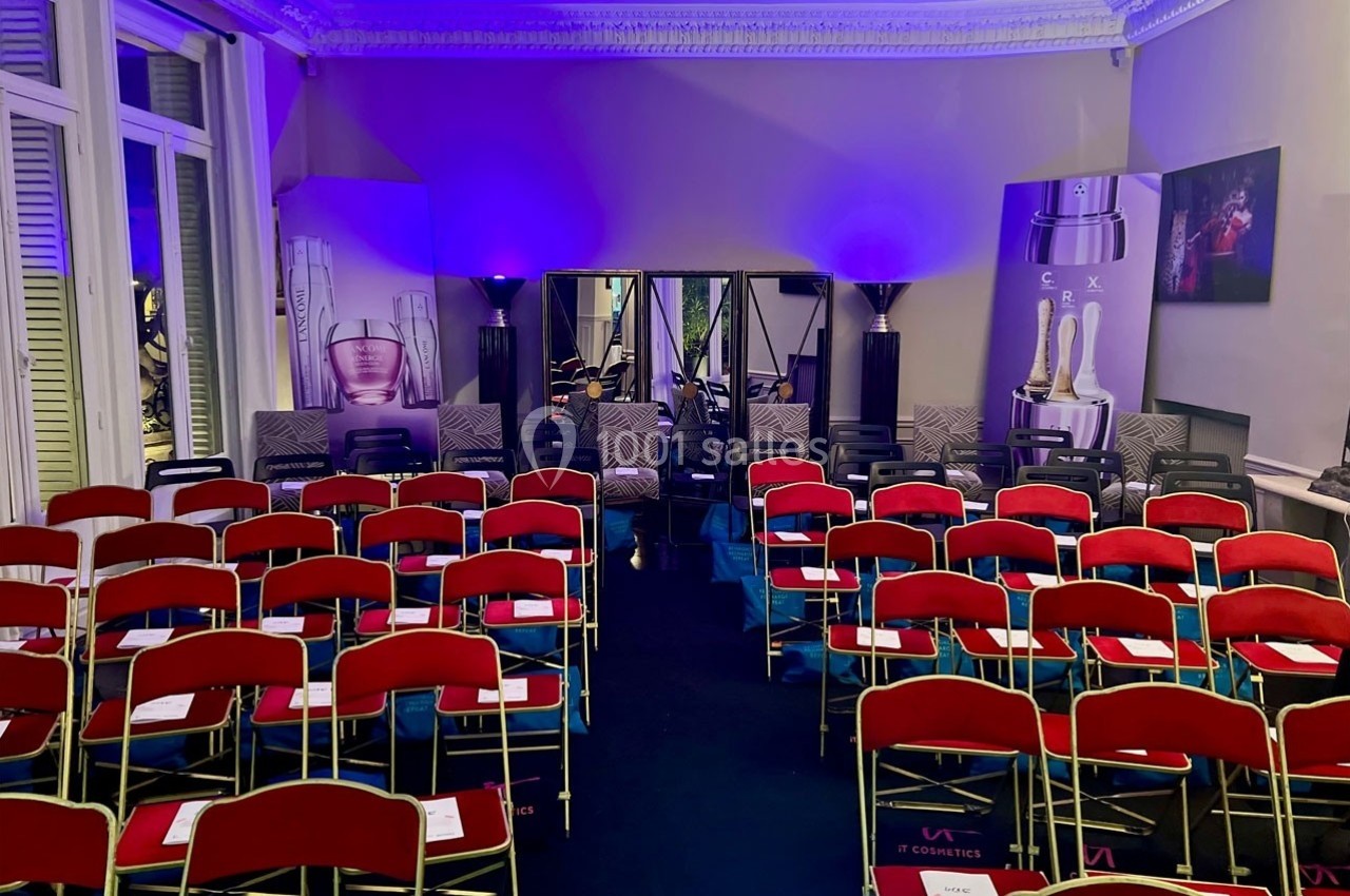 Salle de conférence avec rangées de chaises rouges alignées, éclairage violet et affiches promotionnelles en arrière-plan.