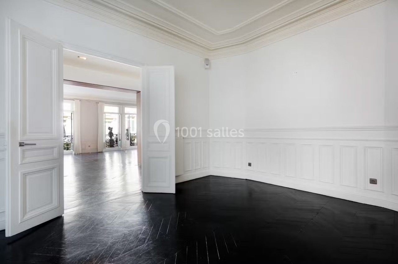 Pièce vide avec parquet sombre, murs blancs ornés de moulures et portes ouvertes sur une autre pièce lumineuse.