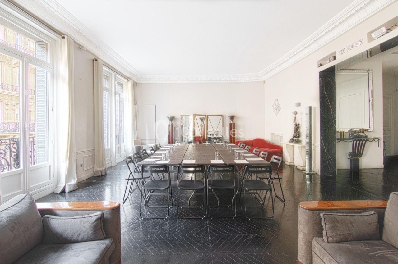 Salle lumineuse avec grande table de réunion, chaises noires, miroirs, canapé rouge et fenêtres donnant sur un balcon.