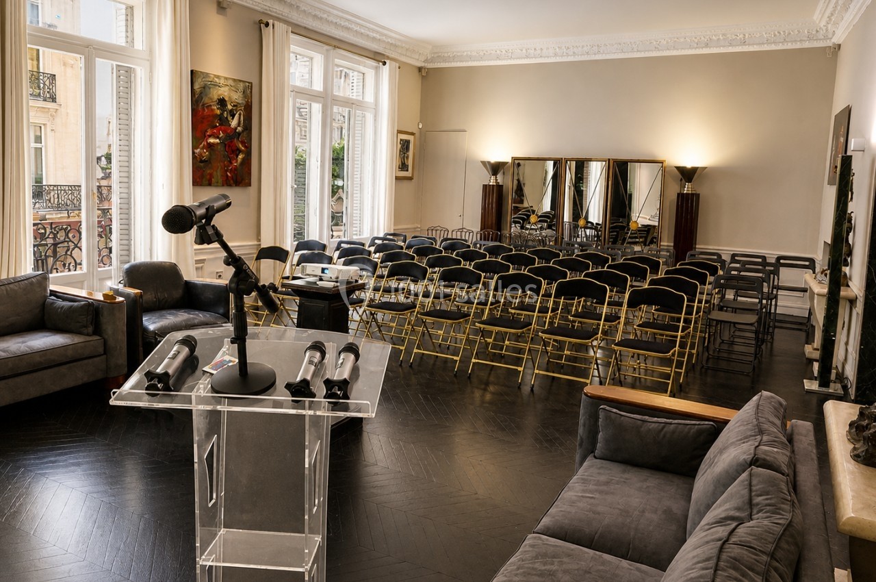 Salle lumineuse aménagée pour une conférence, avec un pupitre, des micros et des rangées de chaises face à des miroirs.