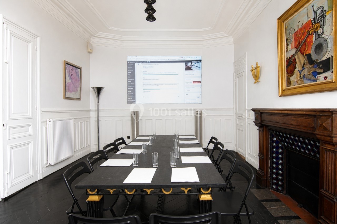 Salle de réunion lumineuse avec une table noire, des chaises pliantes, un écran projeté et des œuvres d'art accrochées.