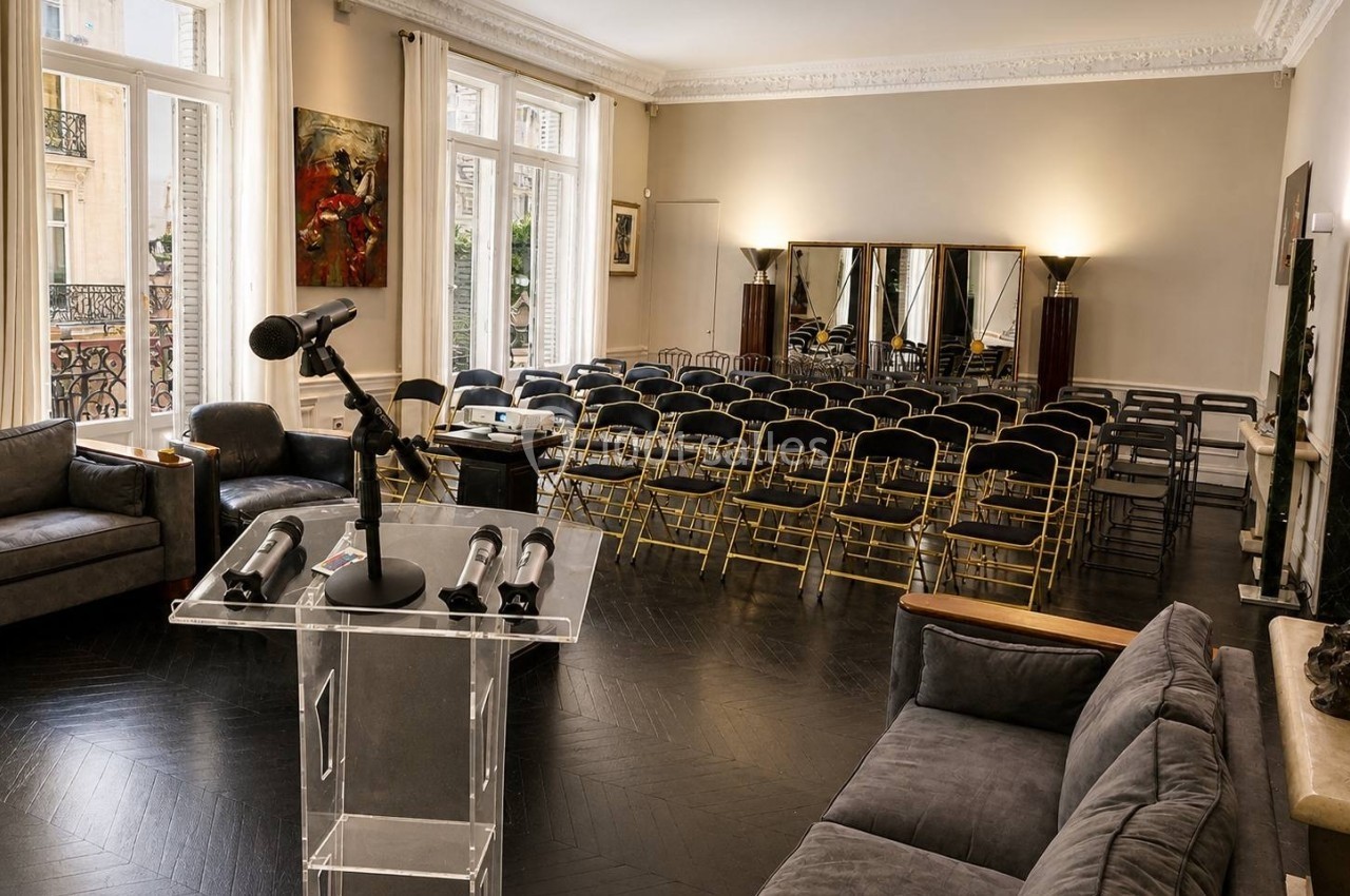 Salle lumineuse aménagée pour une conférence, avec un pupitre transparent, des rangées de chaises et de grandes fenêtres.