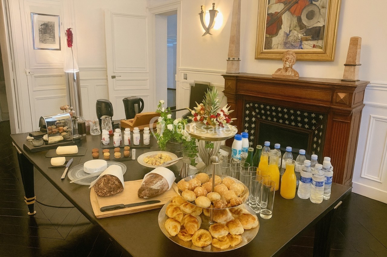 Table dressée avec viennoiseries, pain, boissons, fruits et condiments dans une salle élégante avec cheminée et décorations.