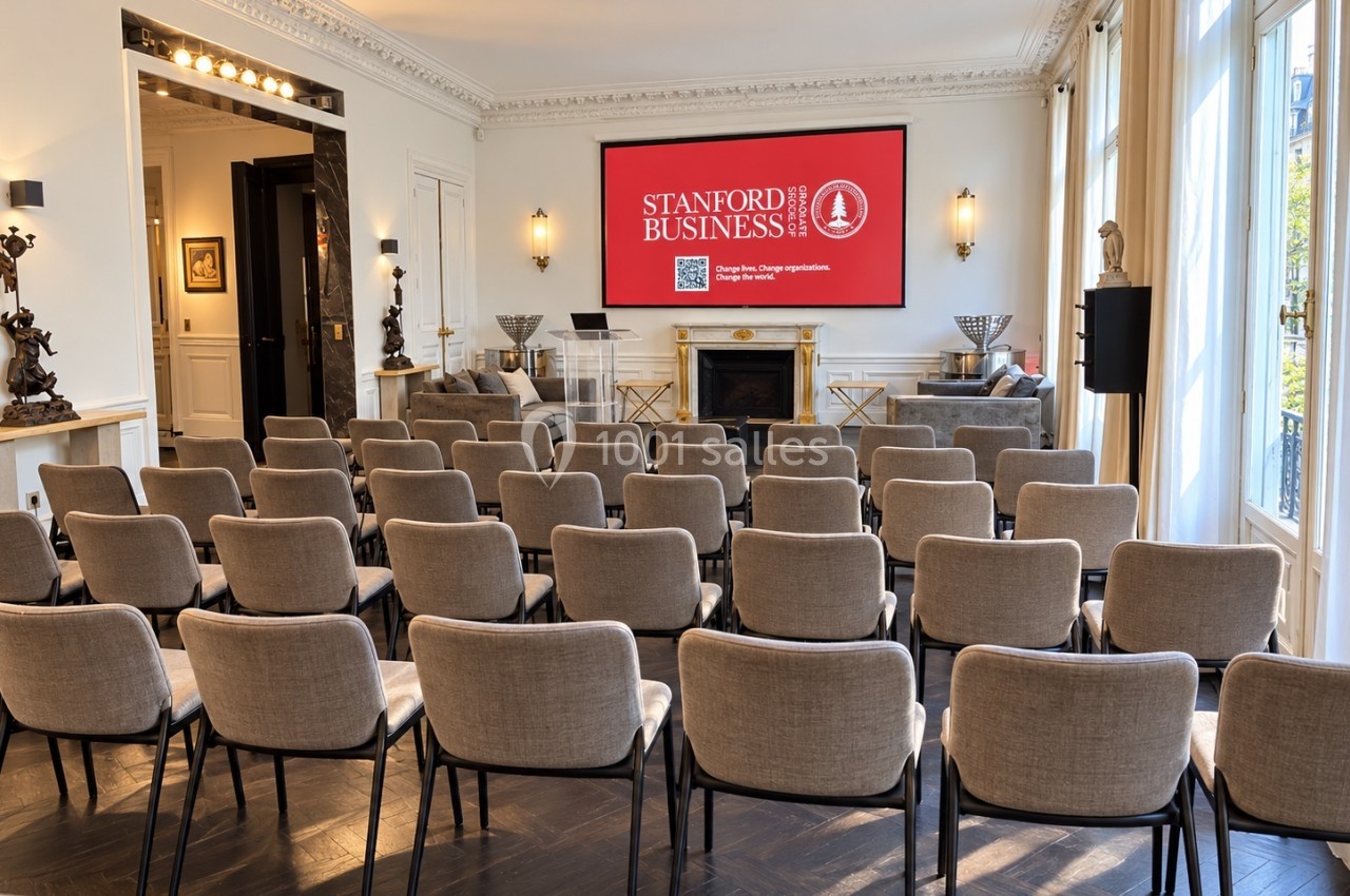 Salle de conférence lumineuse avec des rangées de chaises beige face à un écran affichant ’Stanford Business’.
