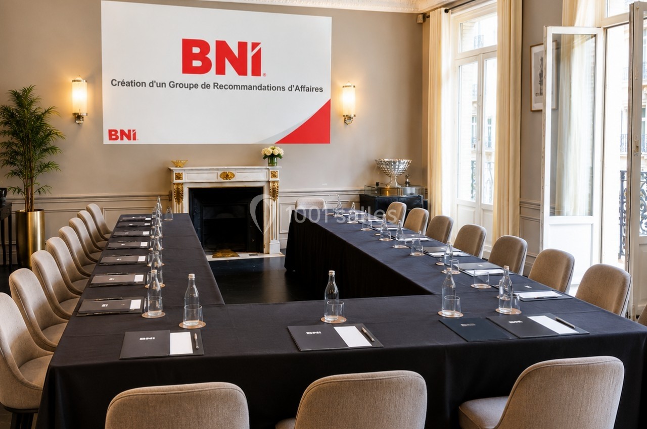Salle de réunion lumineuse avec une grande table en U, chaises beige, bouteilles d'eau et un panneau BNI en fond.