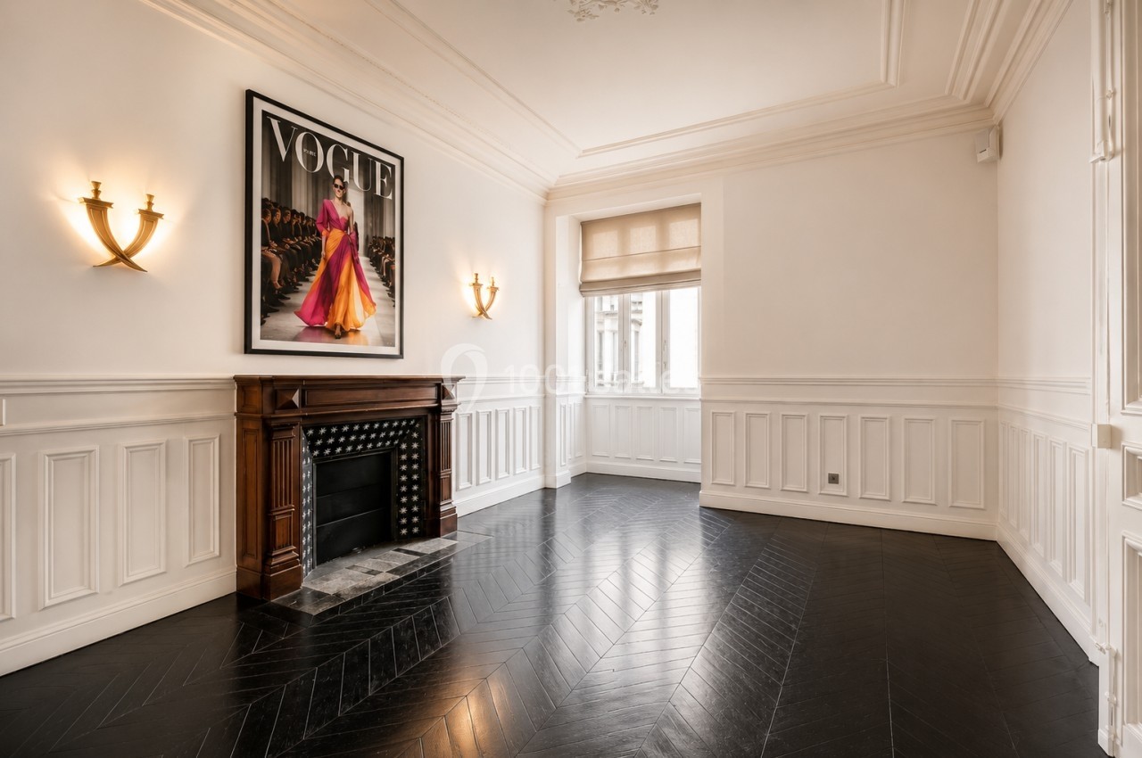 Salle lumineuse avec parquet en chevrons, cheminée encadrée de deux appliques et grande affiche murale.