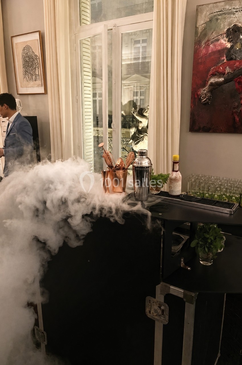 Bar avec fumée, contenant en cuivre rempli de bouteilles, shakers, herbes fraîches et verrerie devant une fenêtre.