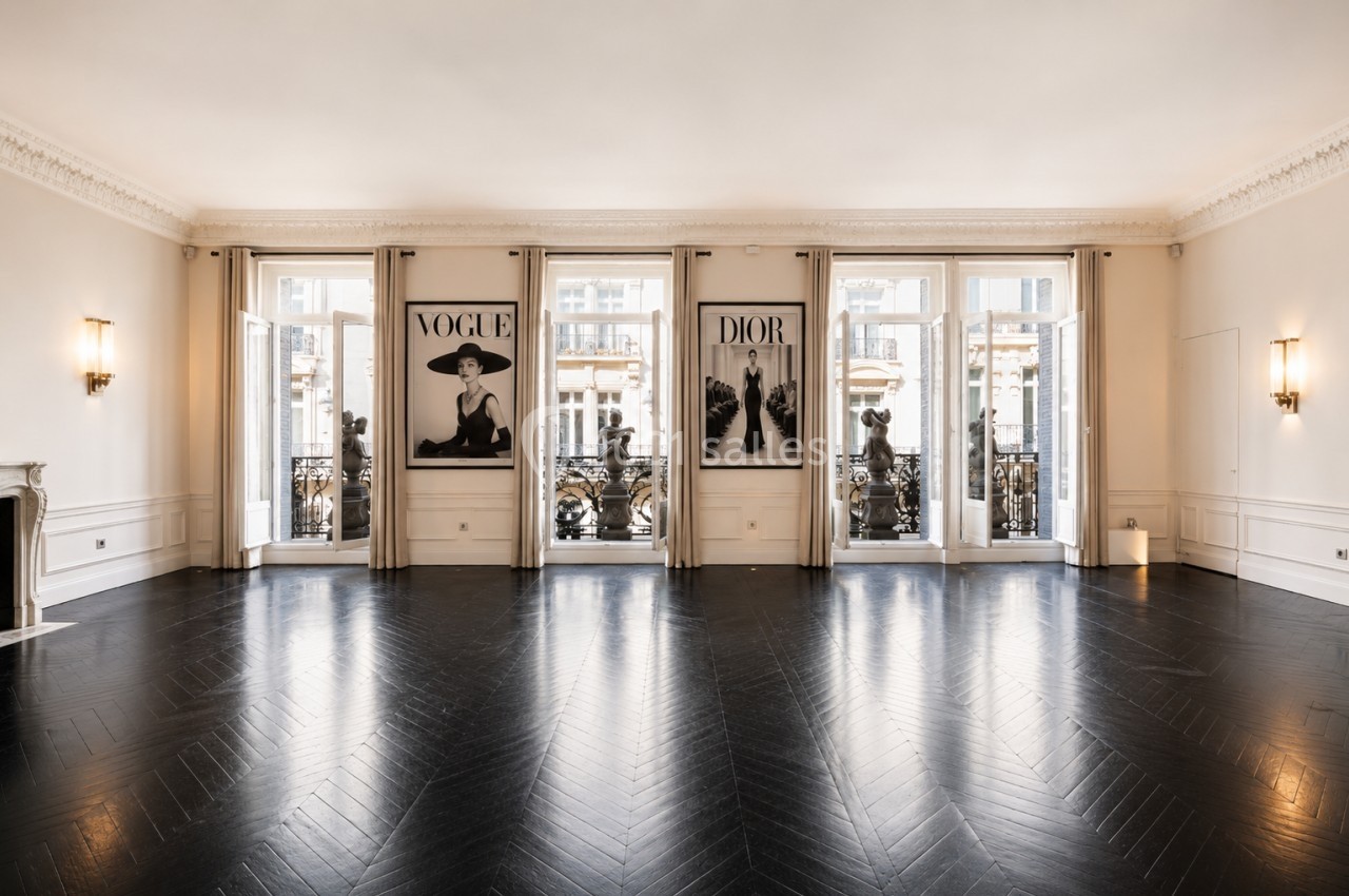 Salle lumineuse avec parquet en chevrons, grandes fenêtres donnant sur un balcon et affiches décoratives au mur.