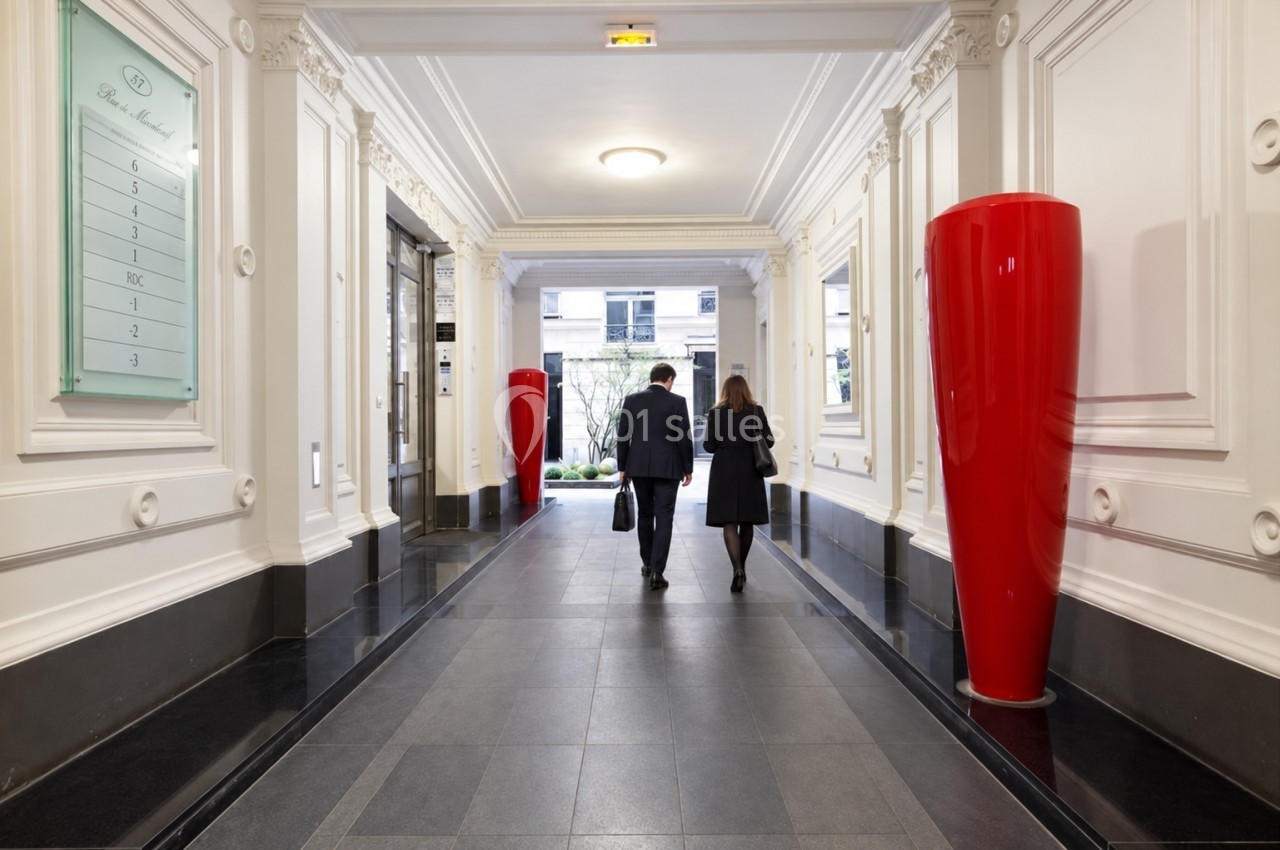 Un homme et une femme marchent dans un couloir élégant avec des colonnes blanches et de grands vases rouges.