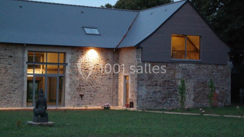 Façade d'une maison en pierre avec de grandes baies vitrées, éclairée en soirée, entourée de pelouse.