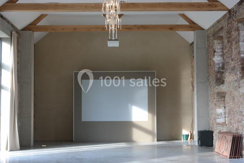 Salle lumineuse avec murs en pierre et bois, écran blanc encadré au centre, plafond avec poutres apparentes et lustre.