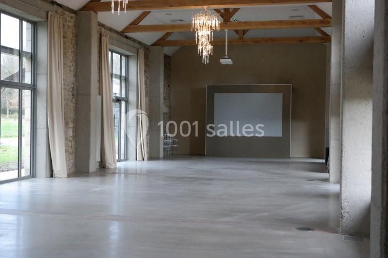 Salle spacieuse avec sol en béton, murs en pierre, poutres apparentes, grandes baies vitrées et un écran blanc.