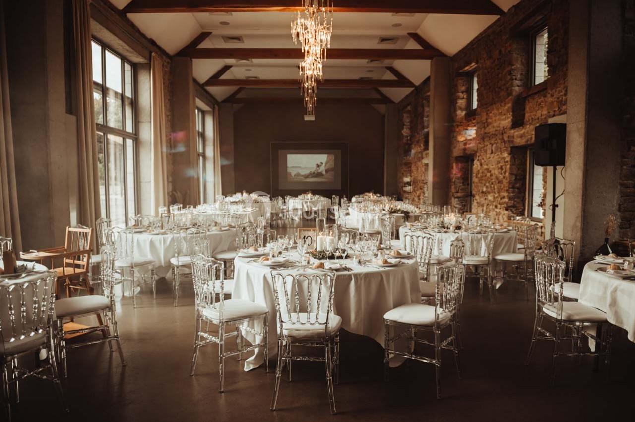 Salle de réception élégante avec tables rondes dressées, chaises transparentes et lustres suspendus.